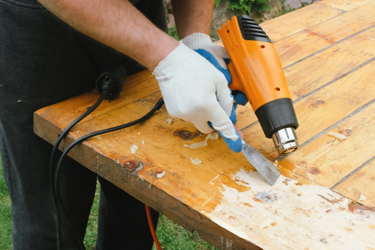 Man removing old varnish from wood using scraper and heat gun.